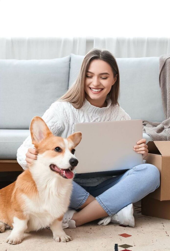 A woman sitting cross-legged on the floor, holding a laptop and petting a cute Corgi that is sitting next to her