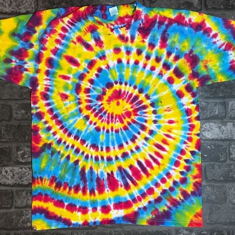 A tie-dye t-shirt in blue, green, pink, white, purple, and orange laid flat against a gray brick background.