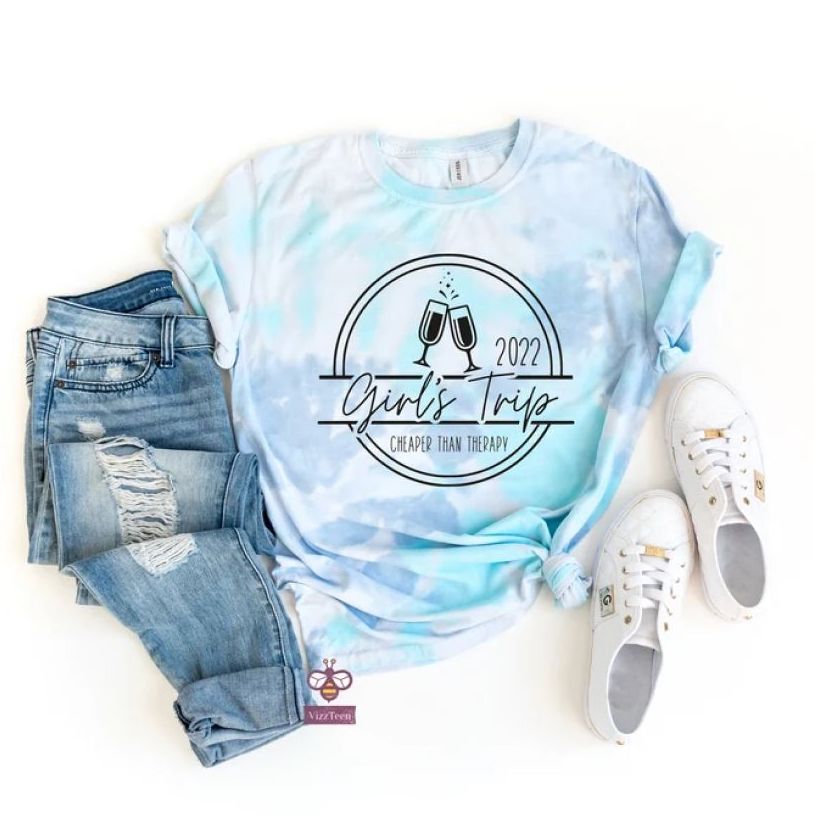 A light blue and purple tie-dye t-shirt with a circular print that says “Girl's trip 2022” and two champagne glasses clinking laid flat beside a pair of blue jeans and white sneakers.