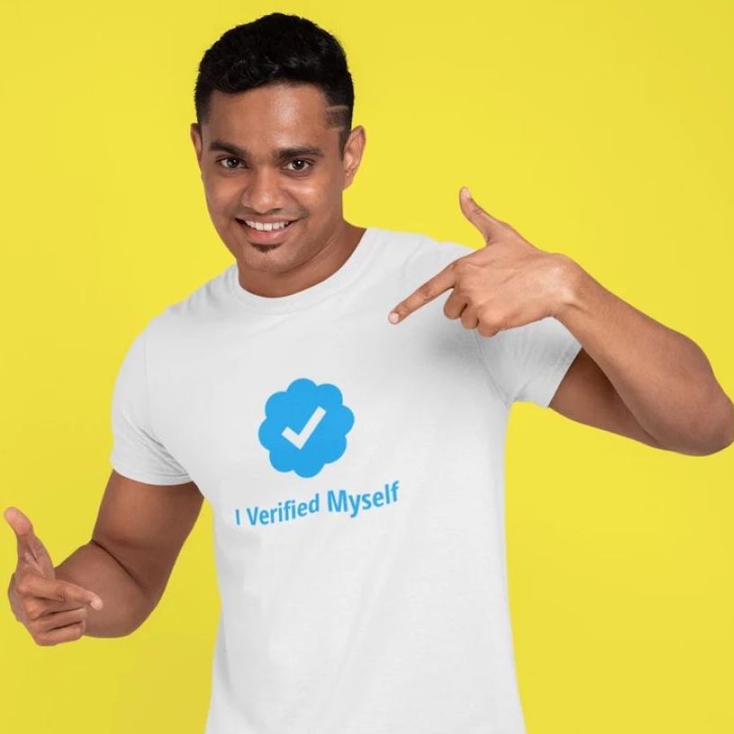 A man on a yellow background wearing a white t-shirt with a print of the Twitter verified icon and the text “I verified myself” in blue.