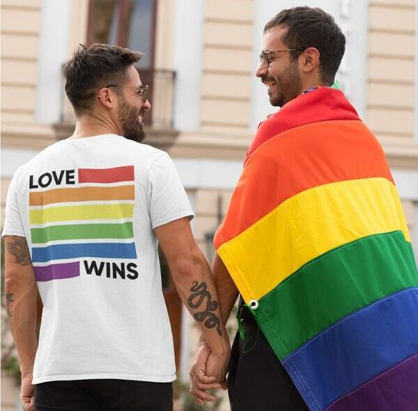 Lifestyle photo of a couple holding hands posing in a rainbow cape and a custom t-shirt with a rainbow design and the slogan “Love Wins” written on the back.