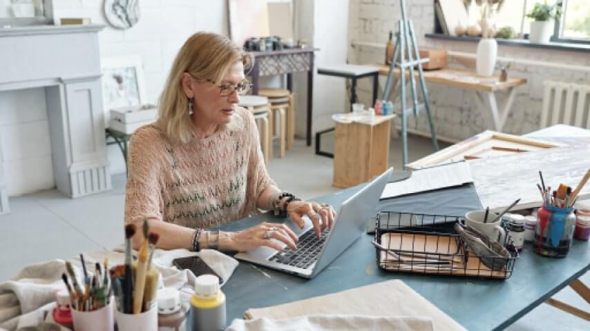 An elderly artist in her studio, using a laptop to learn how to sell art online.