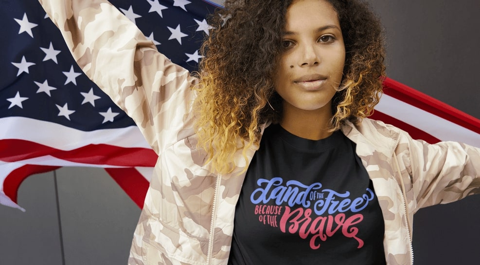 Model wearing a camo jacket and custom t-shirt with the slogan “Land of the Free, Because of the Brave” printed on the chest.