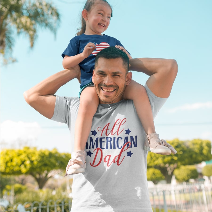Photo of a father holding their child and wearing a t-shirt with the slogan “All American Dad” printed on the chest.
