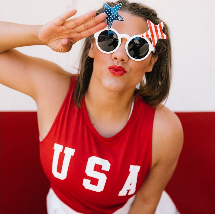 Model in sunglasses peering at the camera and wearing a red tank top with a design reading “USA” printed on the chest.