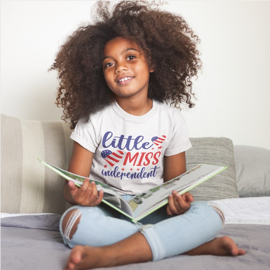 Child posing with an open book and wearing a custom t-shirt with the text “Little miss Independent” printed on the chest.