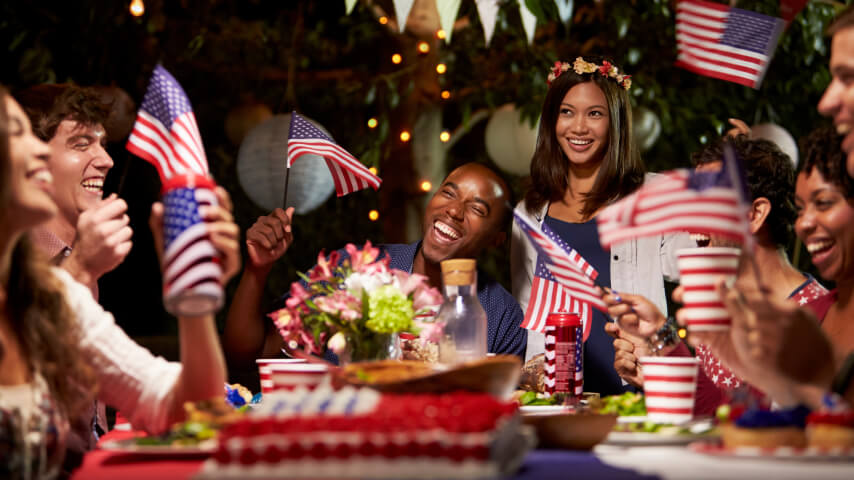 A 4th of July gathering featuring cheerful individuals seated around a table, holding American flags and smiling