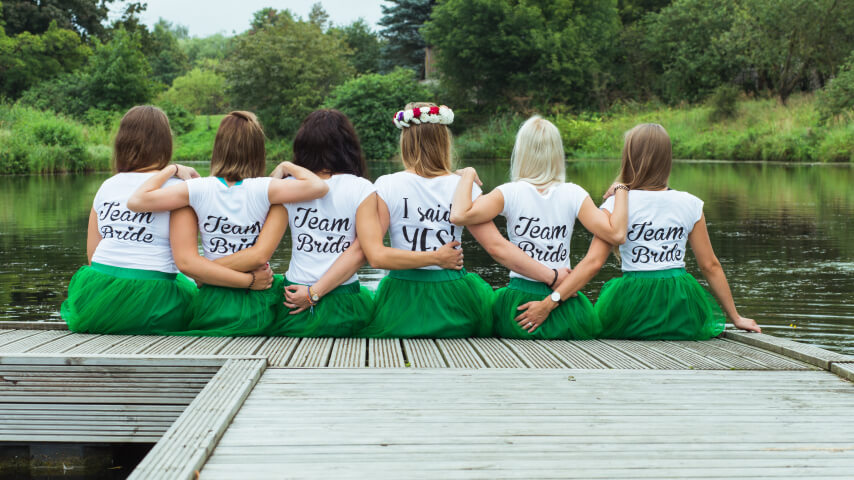 Six bridesmaids wearing matching custom t-shirts and green tulle skirts sit on a footbridge, hugging each other's shoulders.