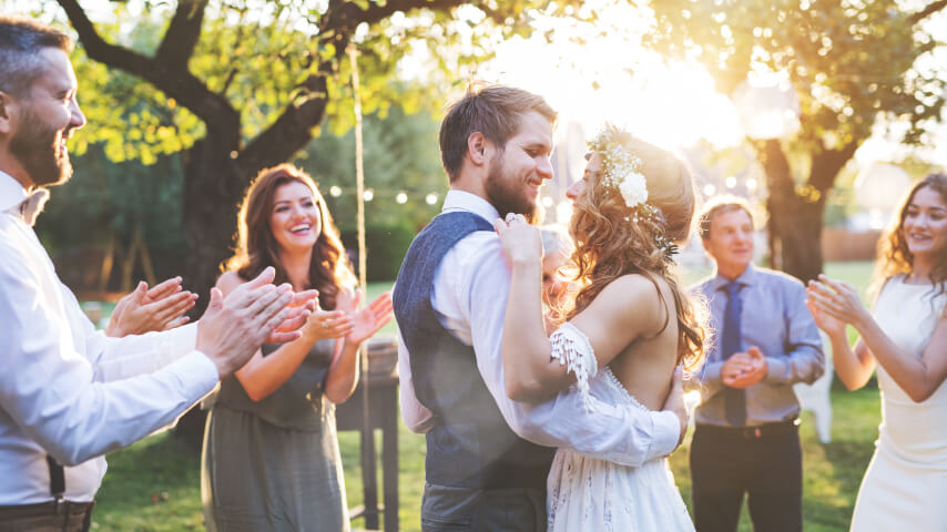 A newlywed couple dancing outdoors, surrounded by beaming guests in the background.
