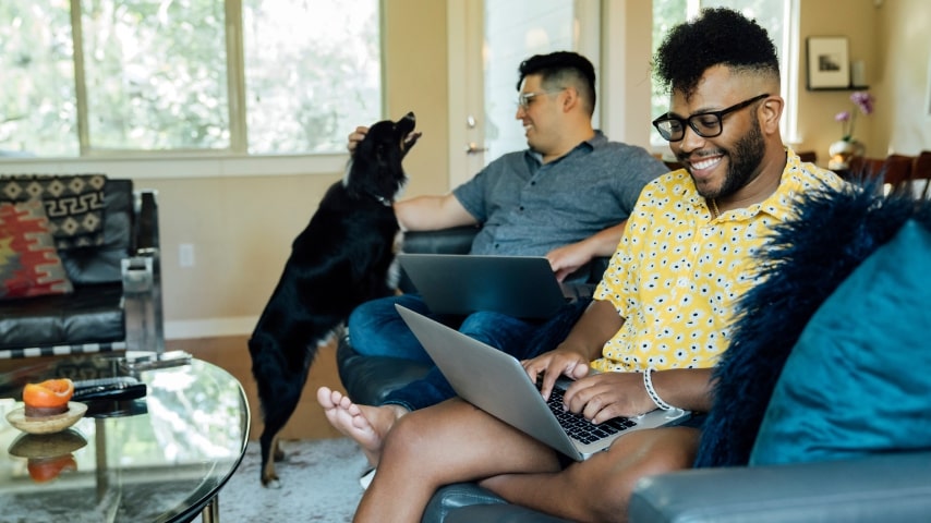 Two men working on their laptops from the comfort of their home, with a dog next to them.