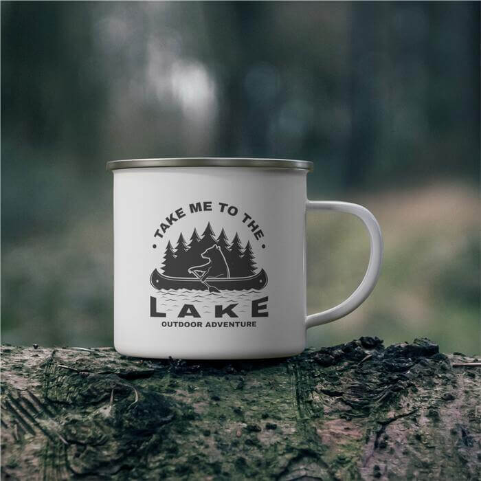 A white enamel mug perched on a tree branch, set against a verdant forest backdrop. The mug showcases an illustration of a bear rowing a boat, encircled by the adventurous call to action, 'take me to the lake'