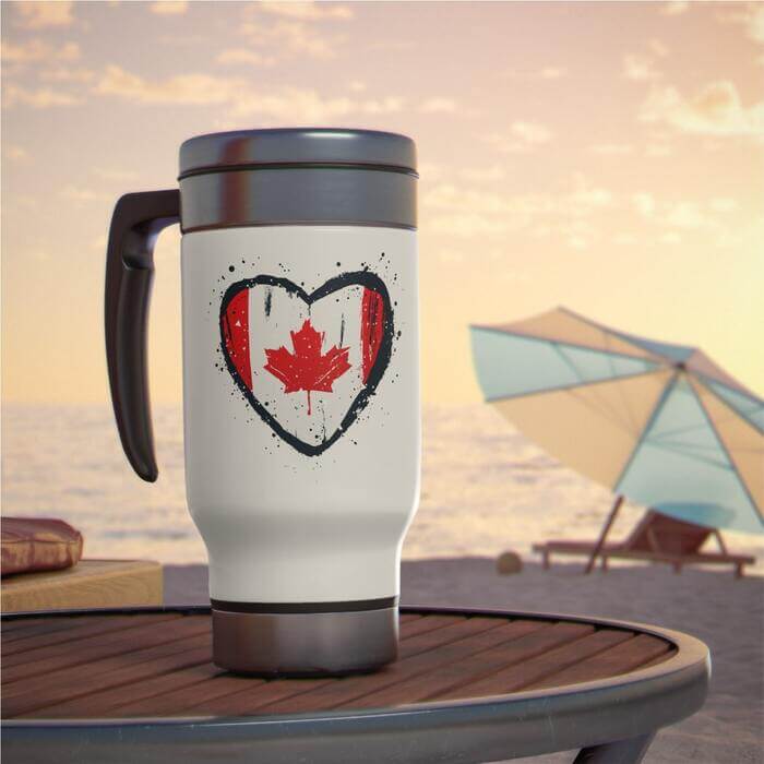 Stainless steel travel mug, decorated with a heart-shaped Canadian flag imprint, casually resting on a table with an inviting beach landscape unfurling in the background