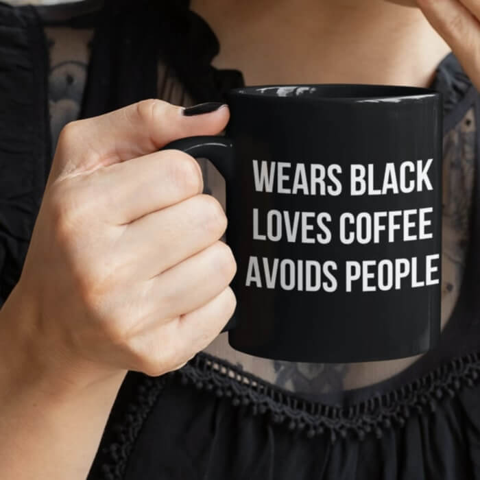 Woman holding a black coffee mug with the text “Wears black, loves coffee, avoids people.”