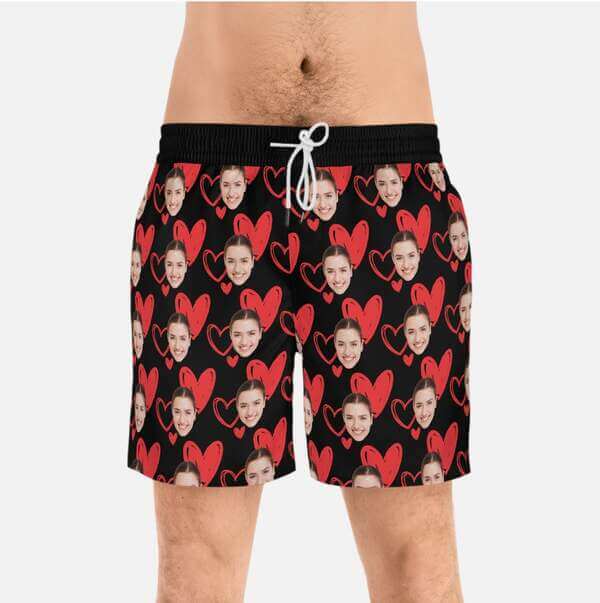Mockup of men's swim trunks with a black base and a repeat pattern of red hearts and a smiling woman's face.