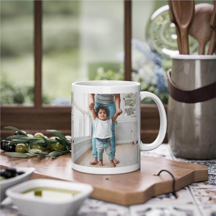 A white mug perfectly placed on a cutting board in a warm kitchen setting. The mug features a heartwarming photo of a cherubic baby taking its first steps, its hands lifted by a supporting parent.