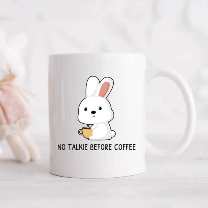 White ceramic mug with a cute, grumpy cartoon bunny holding a coffee cup and the caption “No talkie before coffee.”