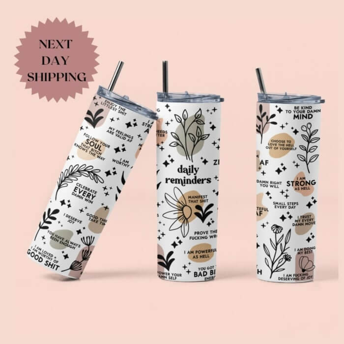 Three tumblers with metal straws and a design of flowers, plants, and “Daily reminders” such as ”Celebrate every damn win,” and more.