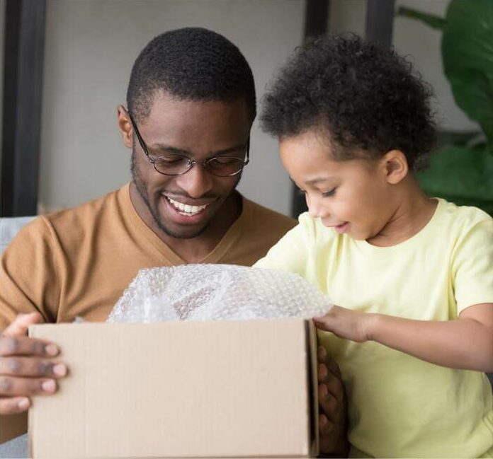 Joyful father and son with dark skin, beaming as they eagerly unbox a surprise from a brown cardboard package