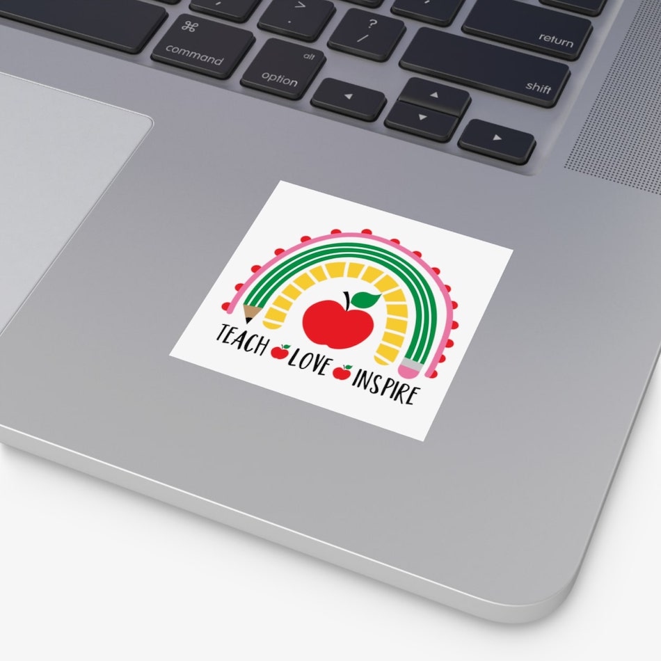 Square sticker on a laptop with a design of a rainbow and apples with the text “Teach. Love. Inspire” underneath.
