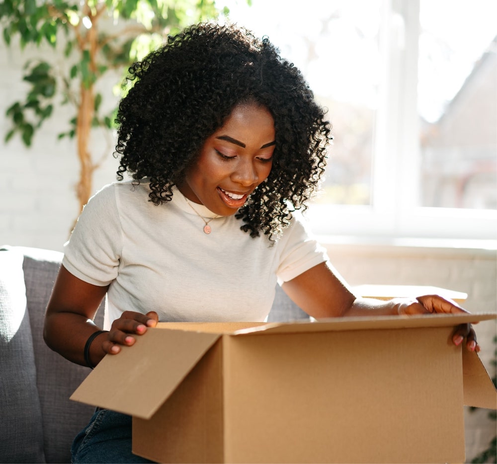 An image of a woman happily unboxing a light-brown cardboard package that has just been delivered.