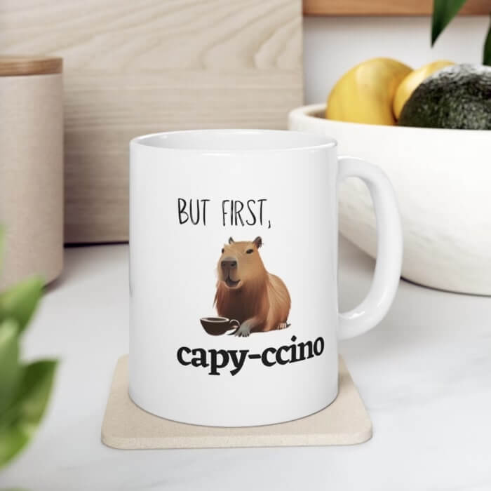 White coffee cup with an image of a capybara and the caption “But first, cappy-ccino.”