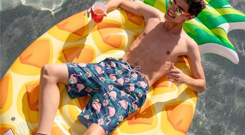 Man with sunglasses on a pool float wearing blue custom swim trunks with a pattern of flowers and flamingos.