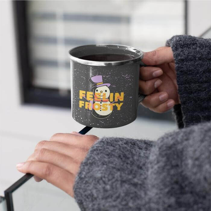 Individual in a cozy grey sweater cradling an enamel mug adorned with a whimsical drawing of a snowman in a vibrant purple top hat, accompanied by the playful, orange-printed phrase 'feelin frosty'