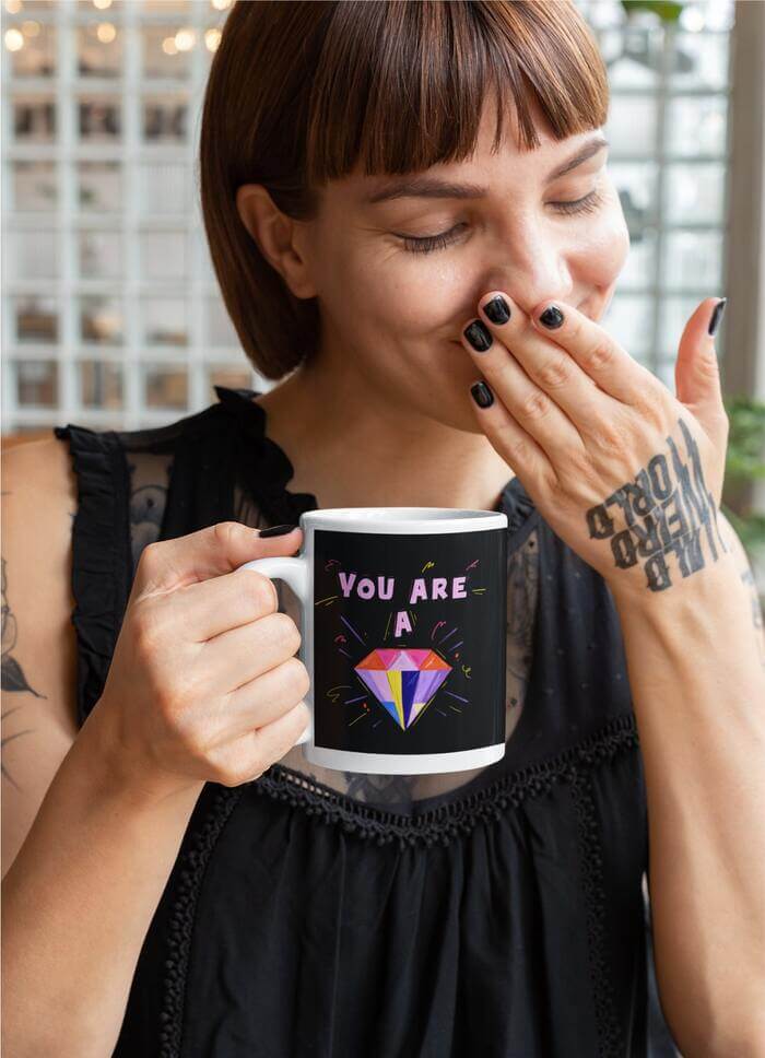 Woman sporting short brown hair with bangs, and an intricate tattoo gracing her hand, holds a mug printed with the empowering phrase 'you are a' alongside a graphic of a diamond.