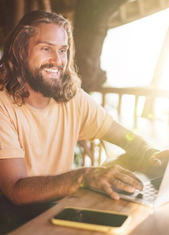 A smiling man with a beard and long hair, working on his laptop in a sunny environment.