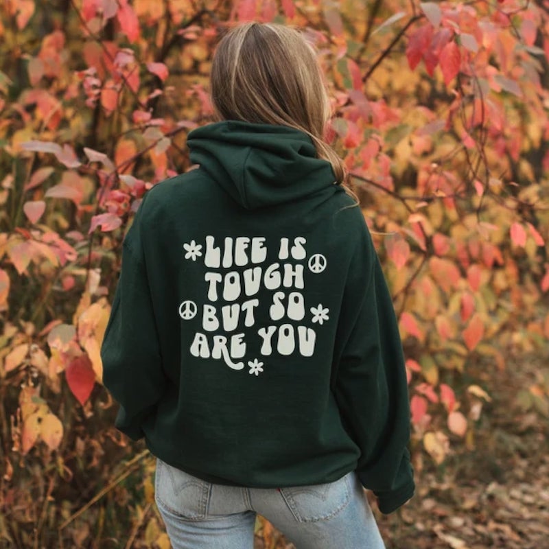 Woman in a dark green hoodie with text on the back saying, “Life is tough but so are you.”