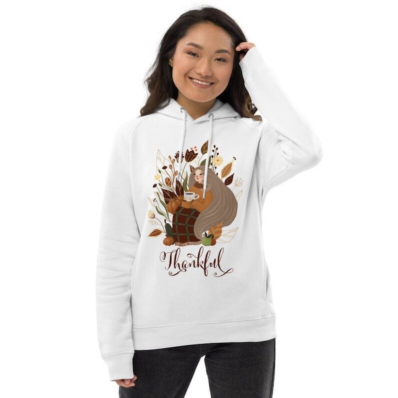 Woman wearing a white hoodie with an autumnal design of a young girl sitting among colorful leaves and flowers, with the text “Thankful” underneath.