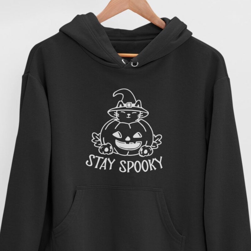 Black hoodie with a white design of a cartoon cat with a witch's hat inside a pumpkin and the text “Stay Spooky” underneath.