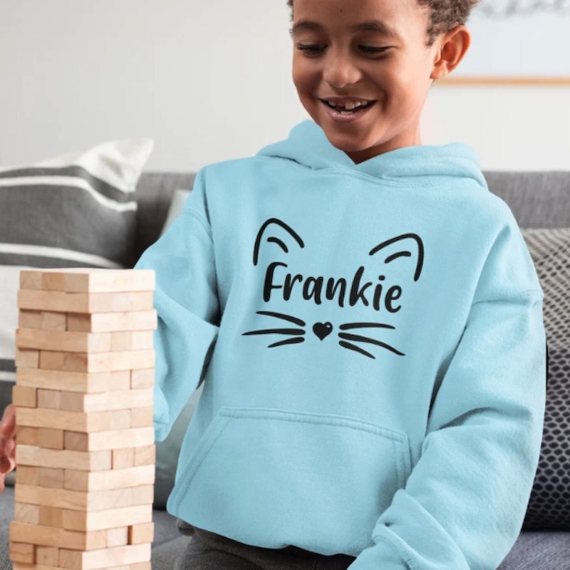 Young boy in a light blue hoodie with a design of cat ears and whiskers and the name “Frankie” printed in the middle.