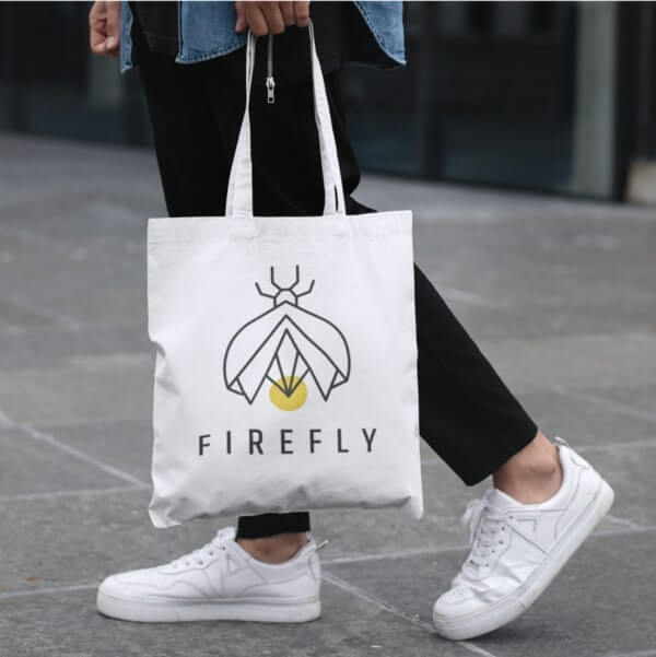 Person holding a white tote bag with a logo of a geometrically stylized bug and the company name “Firefly.”