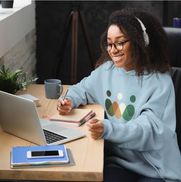 Smiling woman wearing a light blue custom logo hoodie and sitting in front of a laptop at work.