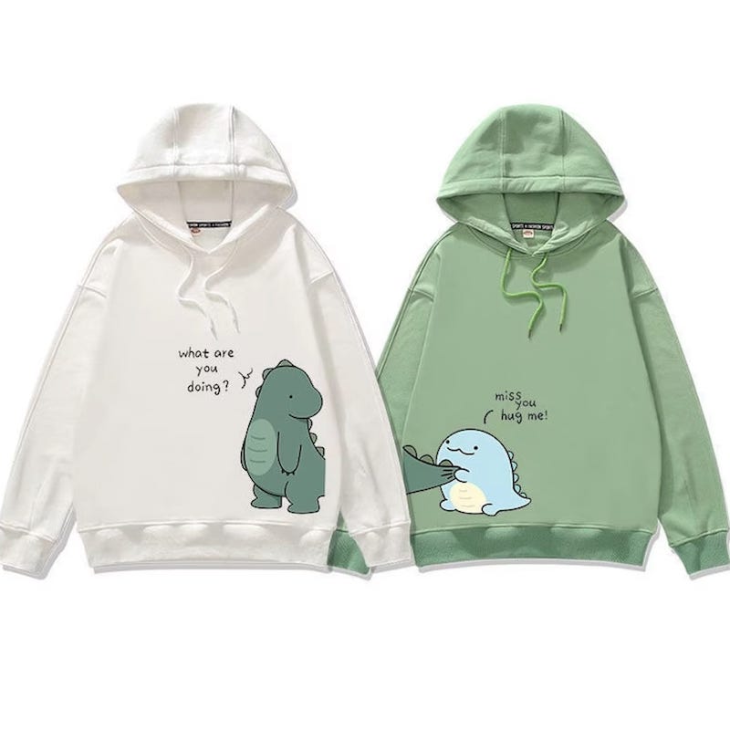 White hoodie with a green dinosaur saying “What are you doing?” next to a green hoodie with a smaller blue dinosaur holding the tail of the first one and saying “miss you, hug me!.”