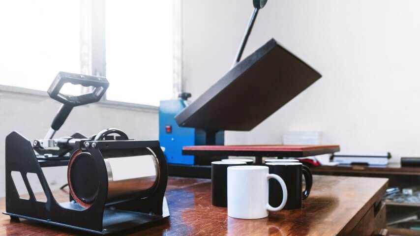 Mug sublimation printer placed on a table next to various black and white mugs.
