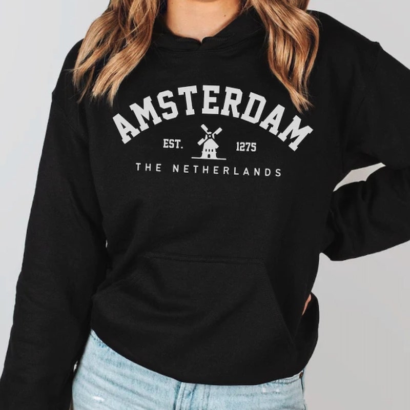 Woman wearing a back hoodie with white text, saying “Amsterdam. Est. 1275. The Netherlands” and a small image of a windmill in the middle.