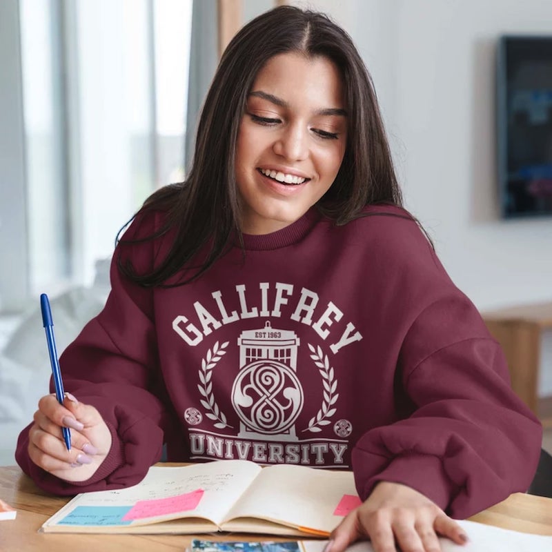 Girl sitting at a desk, wearing a maroon hoodie with a large logo and the text “Gallifrey University.”