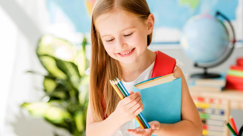 An image of a smiling child in a classroom holding back-to-school supplies: colorful pencils and a book with a blue cover.