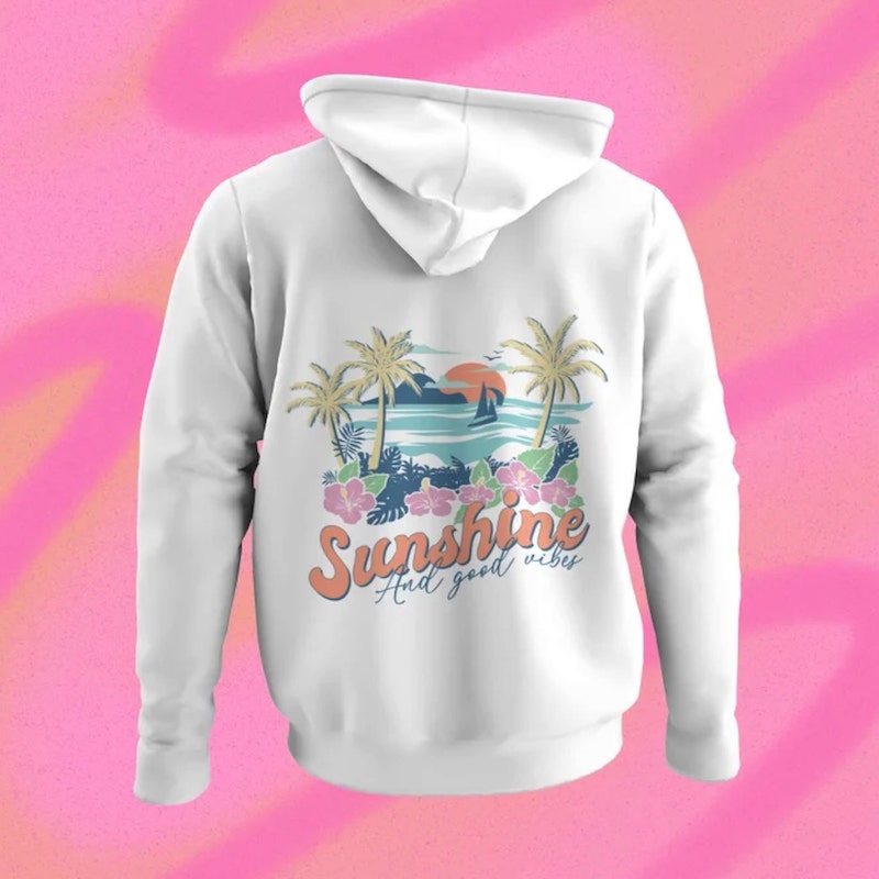 White hoodie with a stylized design of a beach on the back and the text “Sunshine and good vibes” underneath.