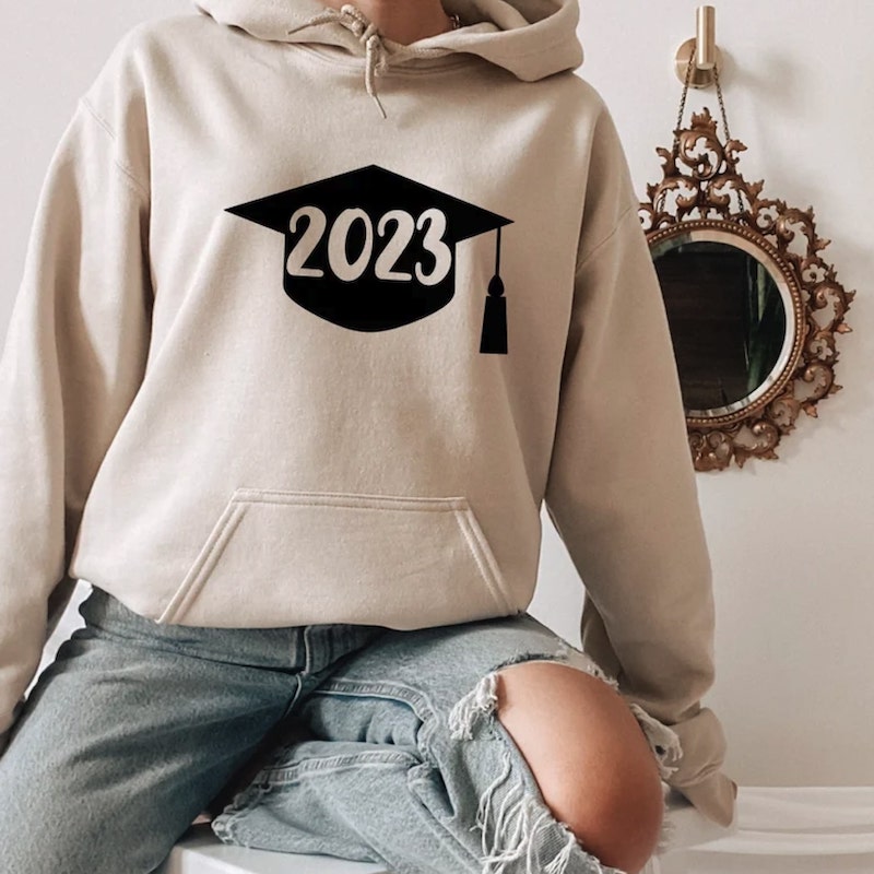 Beige hoodie with a design of a black graduation cap and the year “2023” on it.