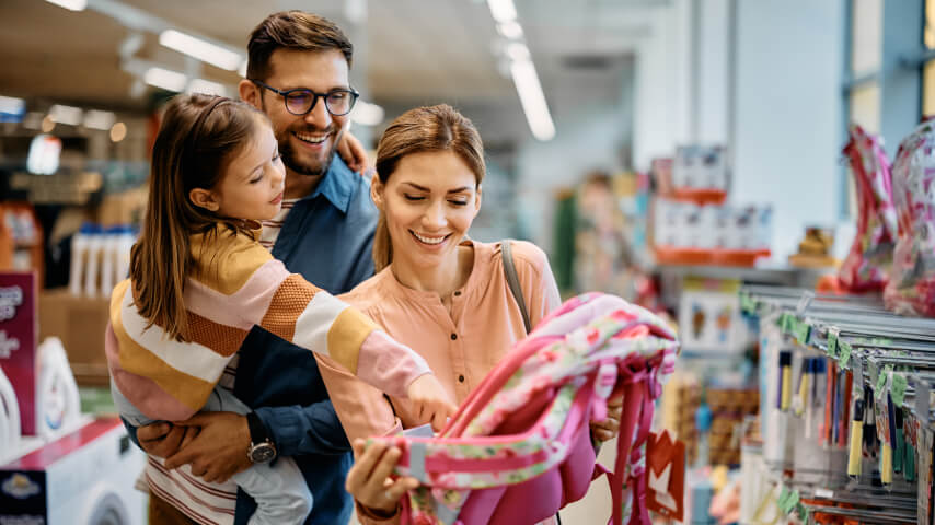 An image of a smiling family (a mother, a father, and their daughter) shopping for back-to-school supplies in a store.