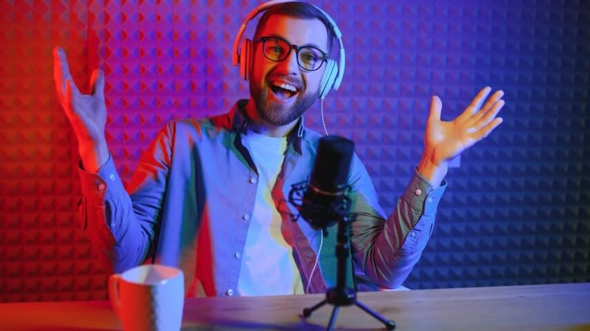 A smiling man with glasses and headphones sitting in a recording studio with neon lighting.
