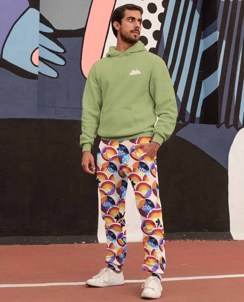 A dark-haired man with a short bear wearing white sneakers, a grayish hoodie, and white joggers with a colorful psychedelic pattern