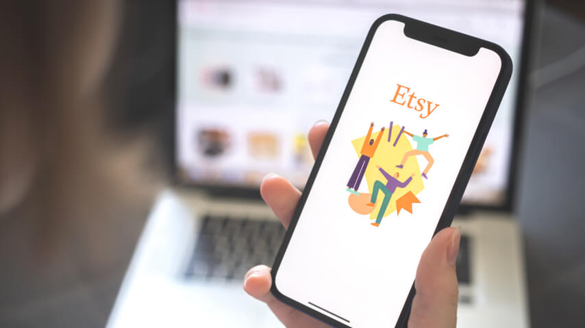 A smartphone displaying an Etsy logo with a blurred laptop in the back; the laptop shows an Etsy page.
