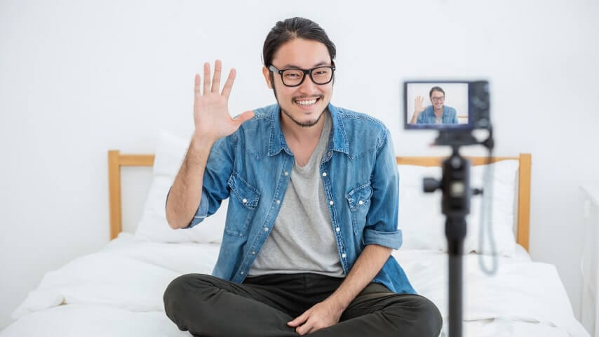 A man waving at a camera while sitting on a bed.