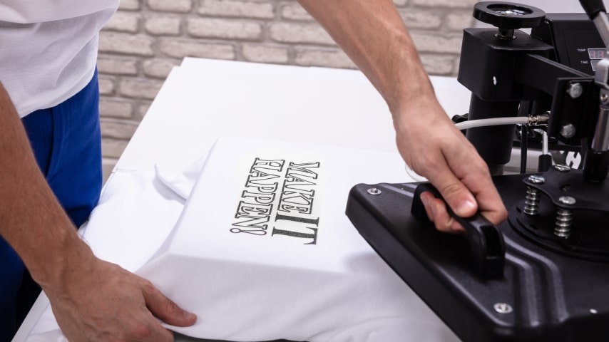 Person using a heat press to print a design that says, “Make It happen” on a white t-shirt.