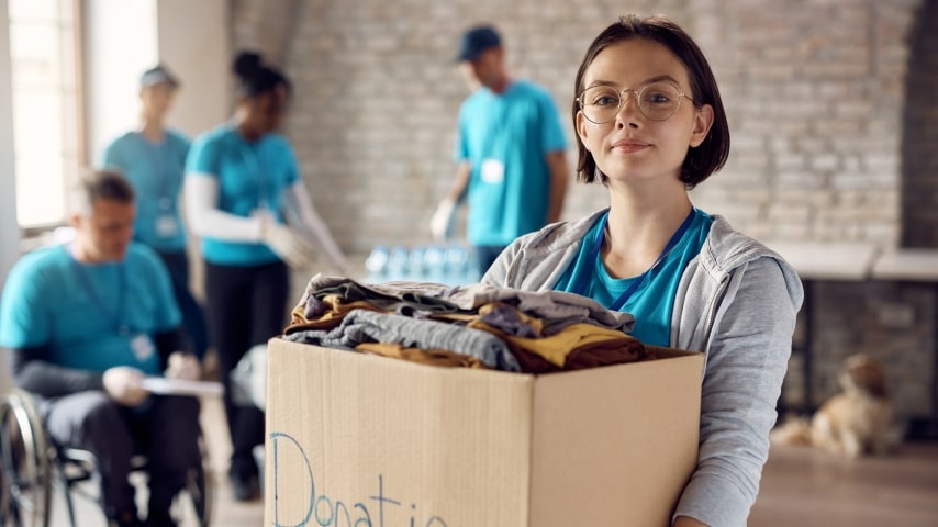 A young woman holding a cardboard box full of clothing with the word “Donations” written on it in sharpie.