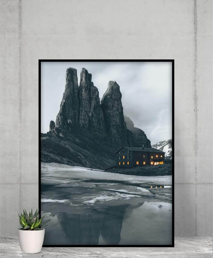 Framed vertical poster of an image of a black house on the beach, with tall dark mountains in the background.
